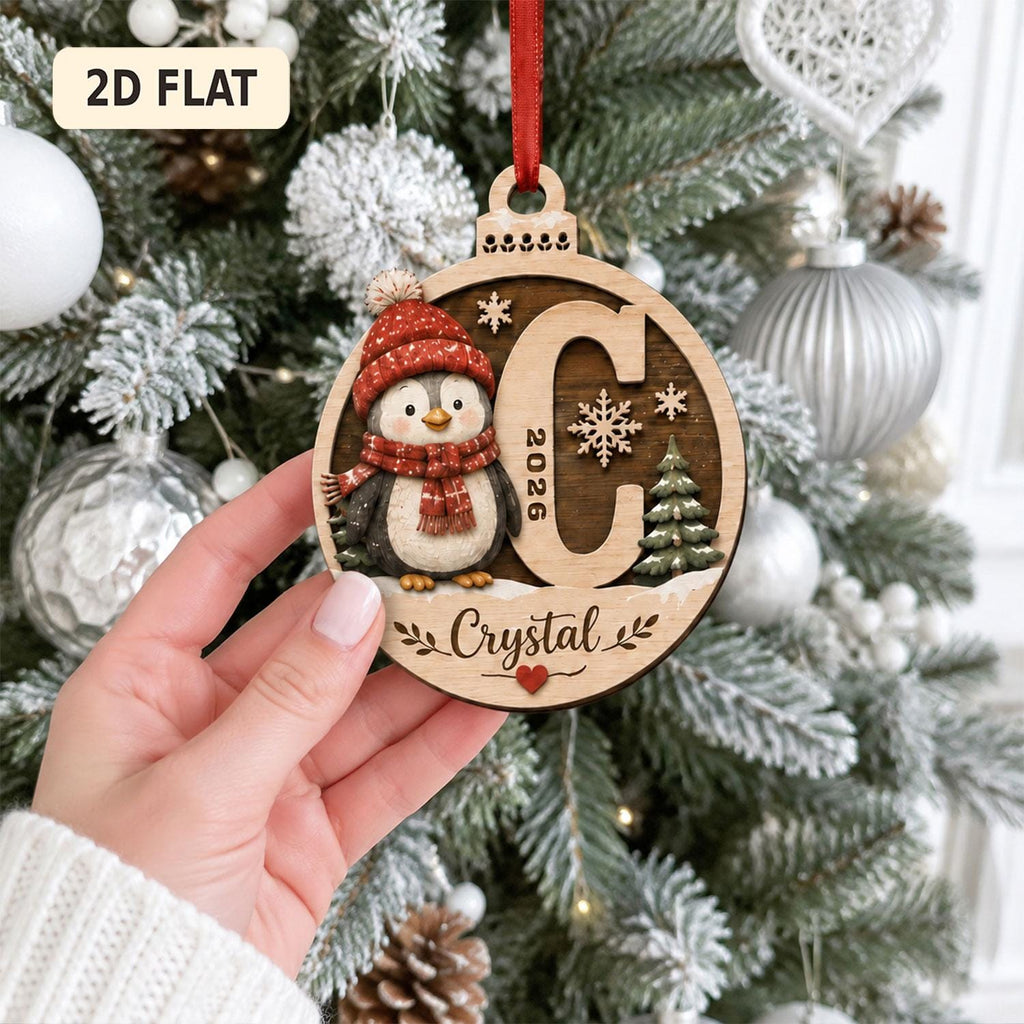 May include: A round wooden Christmas ornament featuring a penguin wearing a red knit hat and scarf, the letter C, the year 2026, a small pine tree, and the name Crystal in script. The ornament hangs from a red ribbon and includes a small red heart.