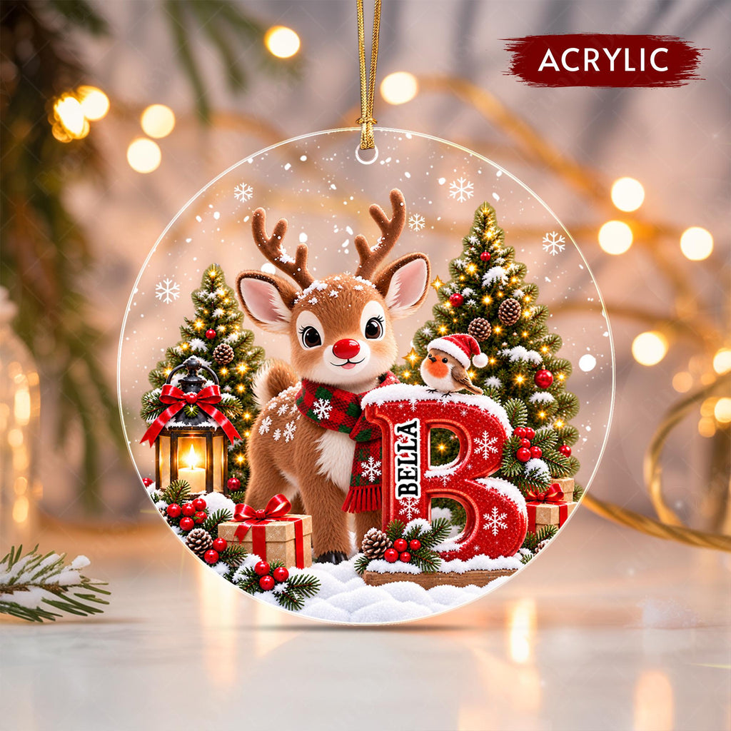 May include: A round acrylic Christmas ornament featuring a cartoon reindeer wearing a red and green plaid scarf, standing next to a large red letter B with the name BELLA printed on it. A small robin, pine trees, a lantern, and gifts are included.