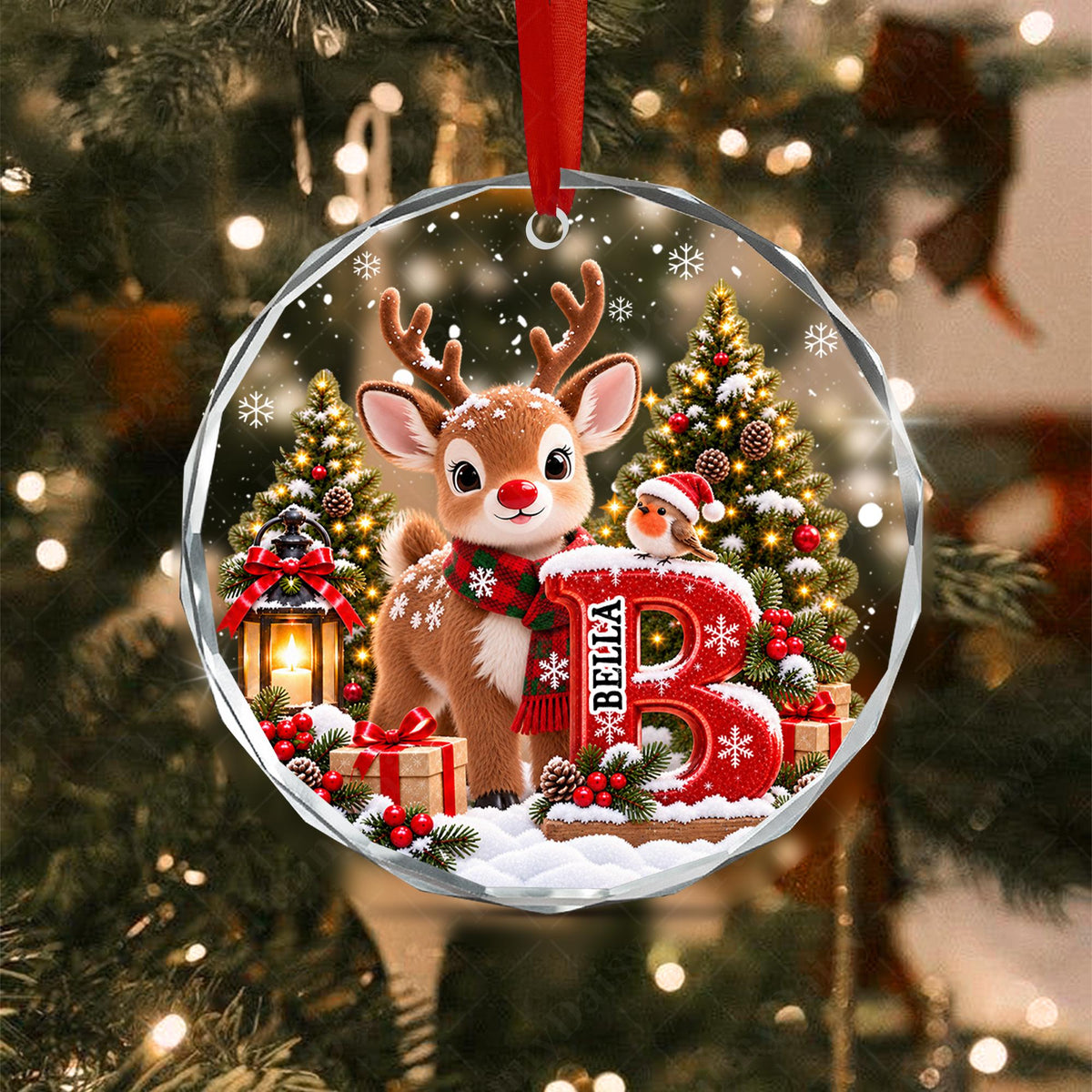 May include: A round, faceted glass Christmas ornament featuring a cartoon reindeer wearing a red and green scarf, a small bird, a lit lantern, and a large red letter B with the name BELLA printed on it, all set against a snowy forest.