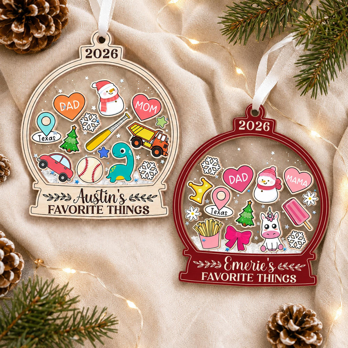 May include: Two snow globe-shaped Christmas ornaments with white ribbons. The left ornament is light wood with '2026' and 'Austin's FAVORITE THINGS' text, featuring icons of a snowman, hearts, a truck, a baseball, and a dinosaur. The right is dark red with '2026' and 'Emerie's FAVORITE THINGS' text, featuring a unicorn, hearts, and fries.