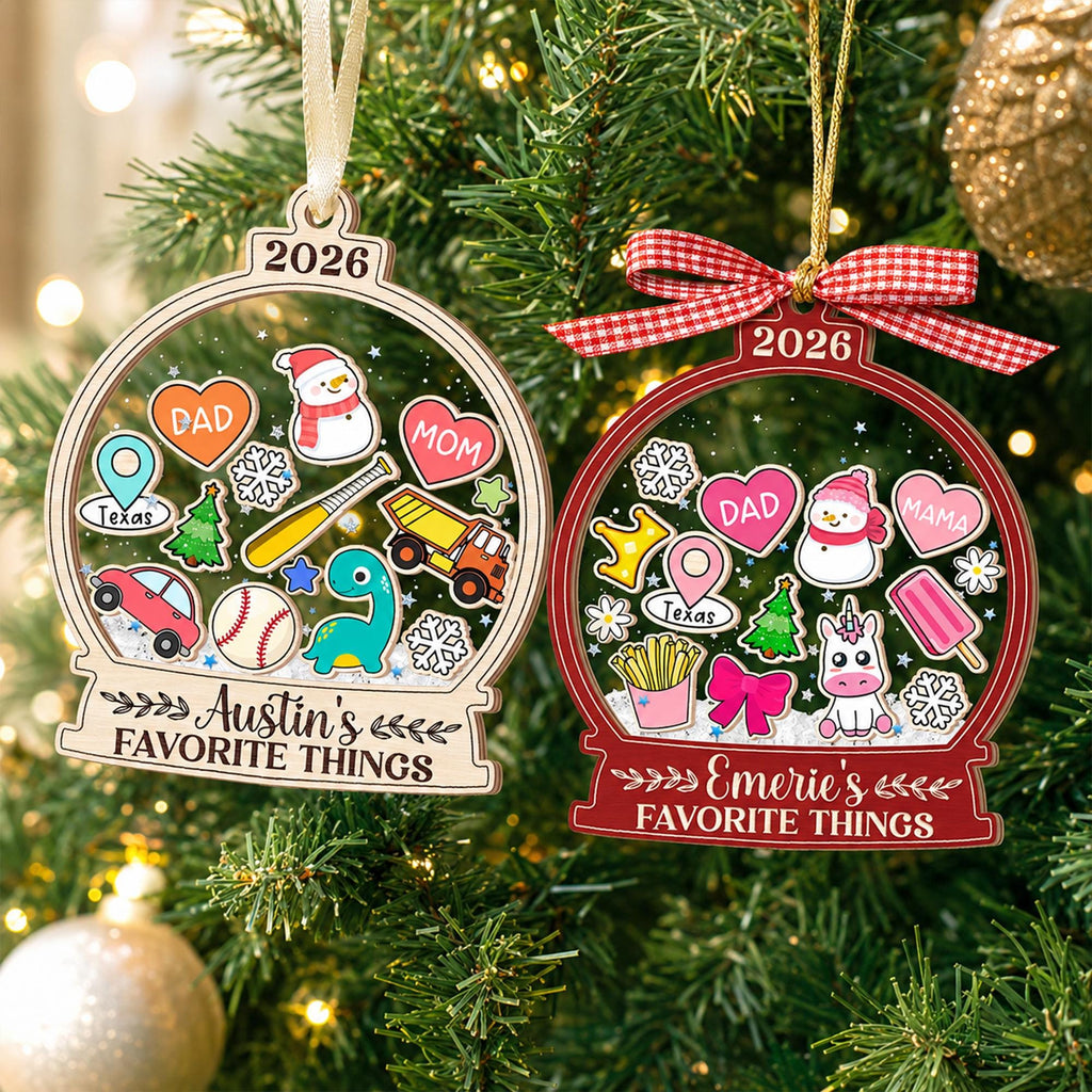 May include: Two globe-shaped Christmas ornaments featuring the year 2026. One is light wood with Austin's FAVORITE THINGS and icons like a snowman, car, and dinosaur. The other is red with Emerie's FAVORITE THINGS and icons like a unicorn and fries.