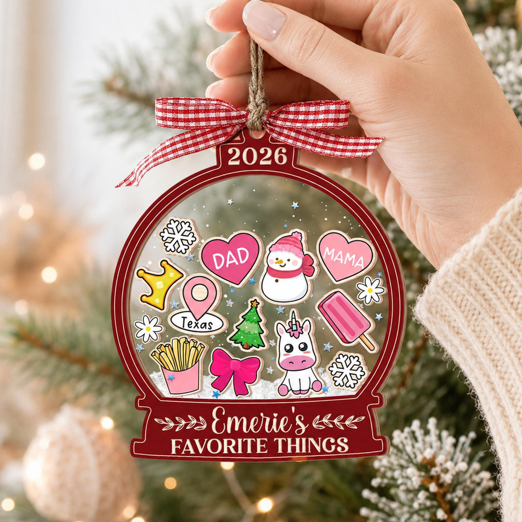 May include: A clear, globe-shaped Christmas ornament with a red border, featuring the text 2026 and Emerie's FAVORITE THINGS. Inside are illustrations of a snowman, unicorn, hearts labeled DAD and MAMA, fries, a bow, and a Texas map pin.