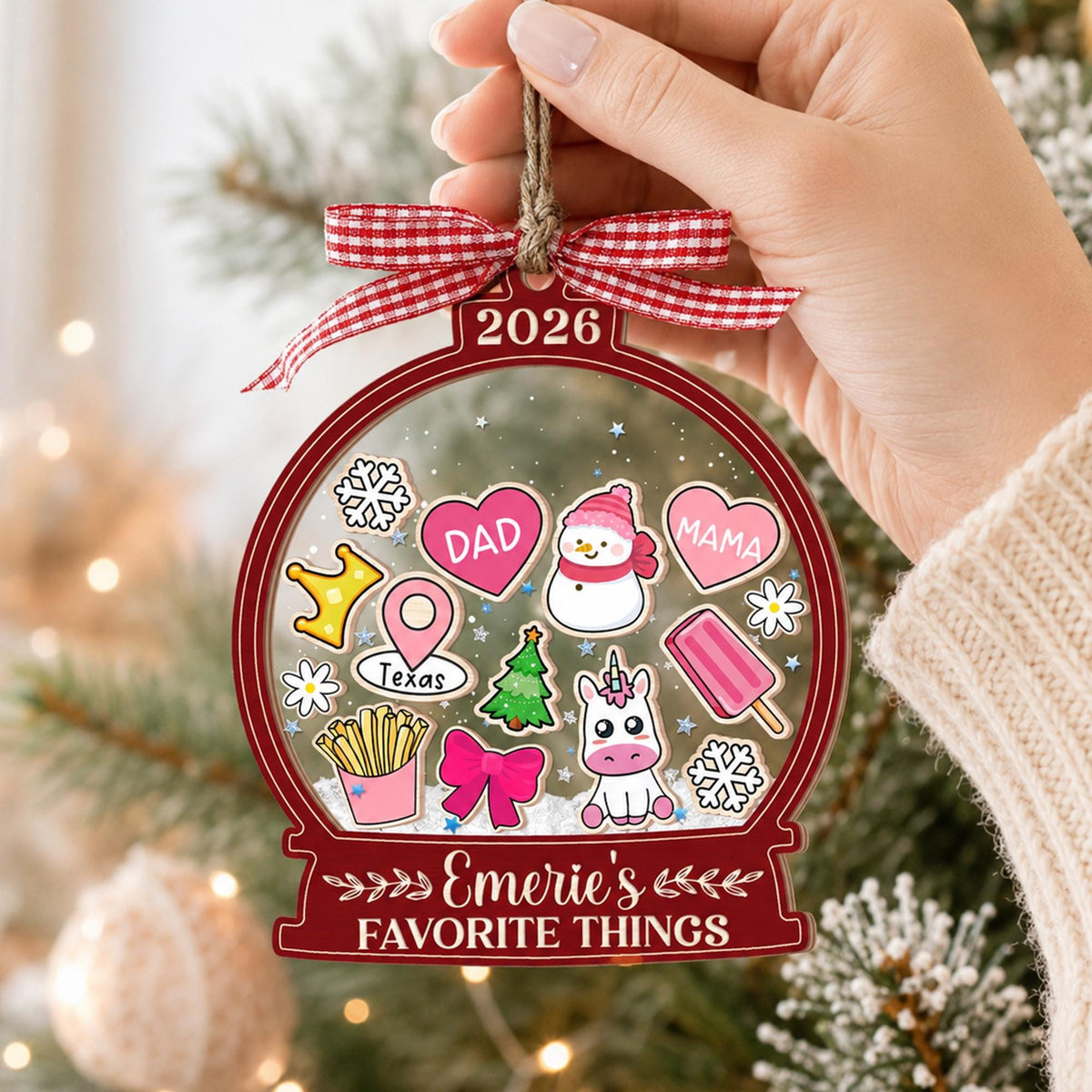 May include: A clear, globe-shaped Christmas ornament with a red border, featuring the text 2026 and Emerie's FAVORITE THINGS. Inside are illustrations of a snowman, unicorn, hearts labeled DAD and MAMA, fries, a bow, and a Texas map pin.
