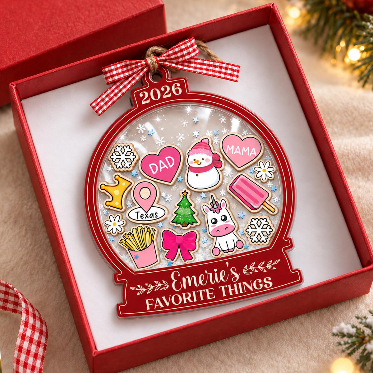 May include: A red snow globe ornament featuring a red and white gingham bow, the year 2026, and the text Emerie's FAVORITE THINGS. Inside are various icons including a snowman, unicorn, fries, hearts labeled DAD and MAMA, and a Texas map pin.