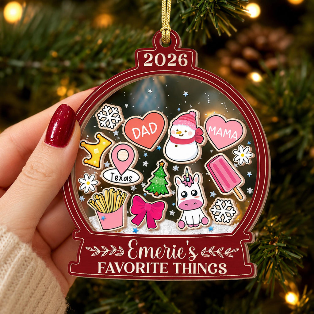 May include: A clear, globe-shaped Christmas ornament with a dark red border, featuring the year 2026 at the top and the text Emerie's FAVORITE THINGS at the bottom. Inside are colorful icons including a snowman, unicorn, hearts labeled DAD and MAMA, a Texas map pin, fries, and snowflakes.