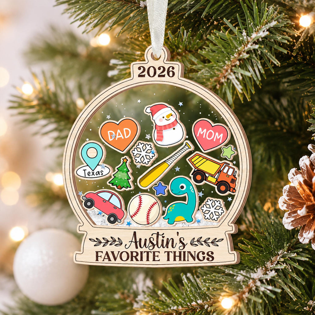 May include: A wooden snow globe ornament featuring the year 2026 at the top and the text Austin's FAVORITE THINGS at the bottom. The center contains colorful illustrations of a snowman, hearts labeled DAD and MOM, a Texas map pin, a baseball bat, a dump truck, a car, a dinosaur, a Christmas tree, snowflakes, and a baseball.