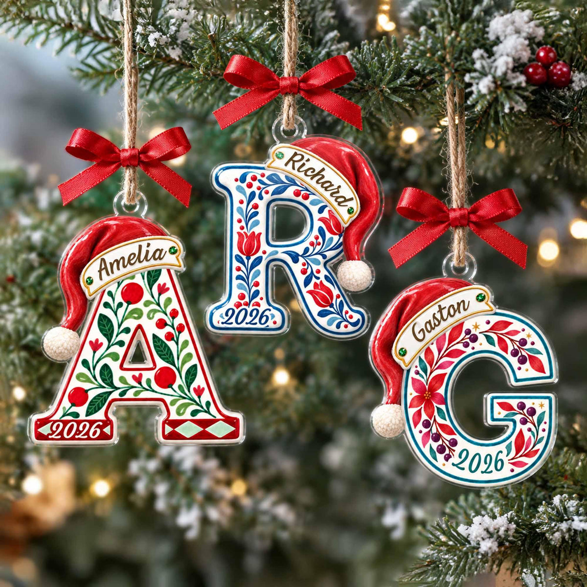May include: Three clear acrylic letter ornaments shaped as A, R, and G, each topped with a red Santa hat and a red satin bow. The letters feature colorful floral patterns, the names Amelia, Richard, and Gaston, and the year 2026 printed on them.