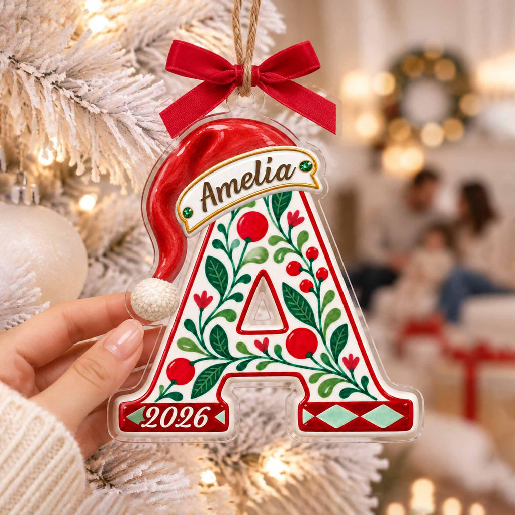 May include: A clear acrylic Christmas ornament shaped like the letter A, topped with a red ribbon bow and a red Santa hat. The letter features green holly leaves, red berries, the name Amelia, and the year 2026 printed in red and green.