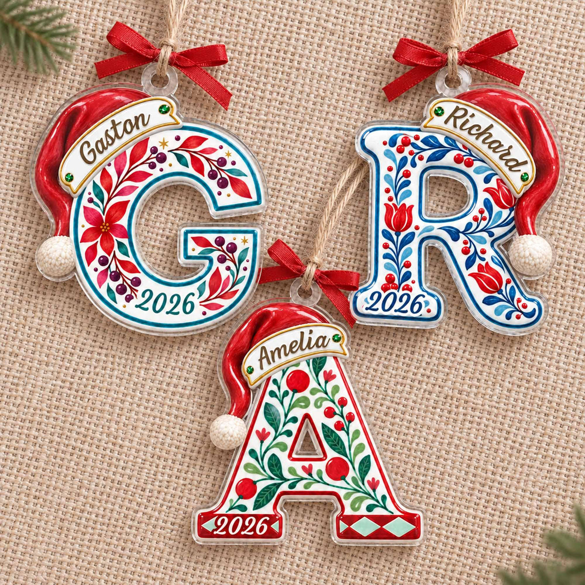 May include: Three clear acrylic letter ornaments shaped as G, R, and A, each topped with a red Santa hat and a red ribbon bow. The letters feature colorful floral patterns, the names Gaston, Richard, and Amelia, and the year 2026 printed on them.