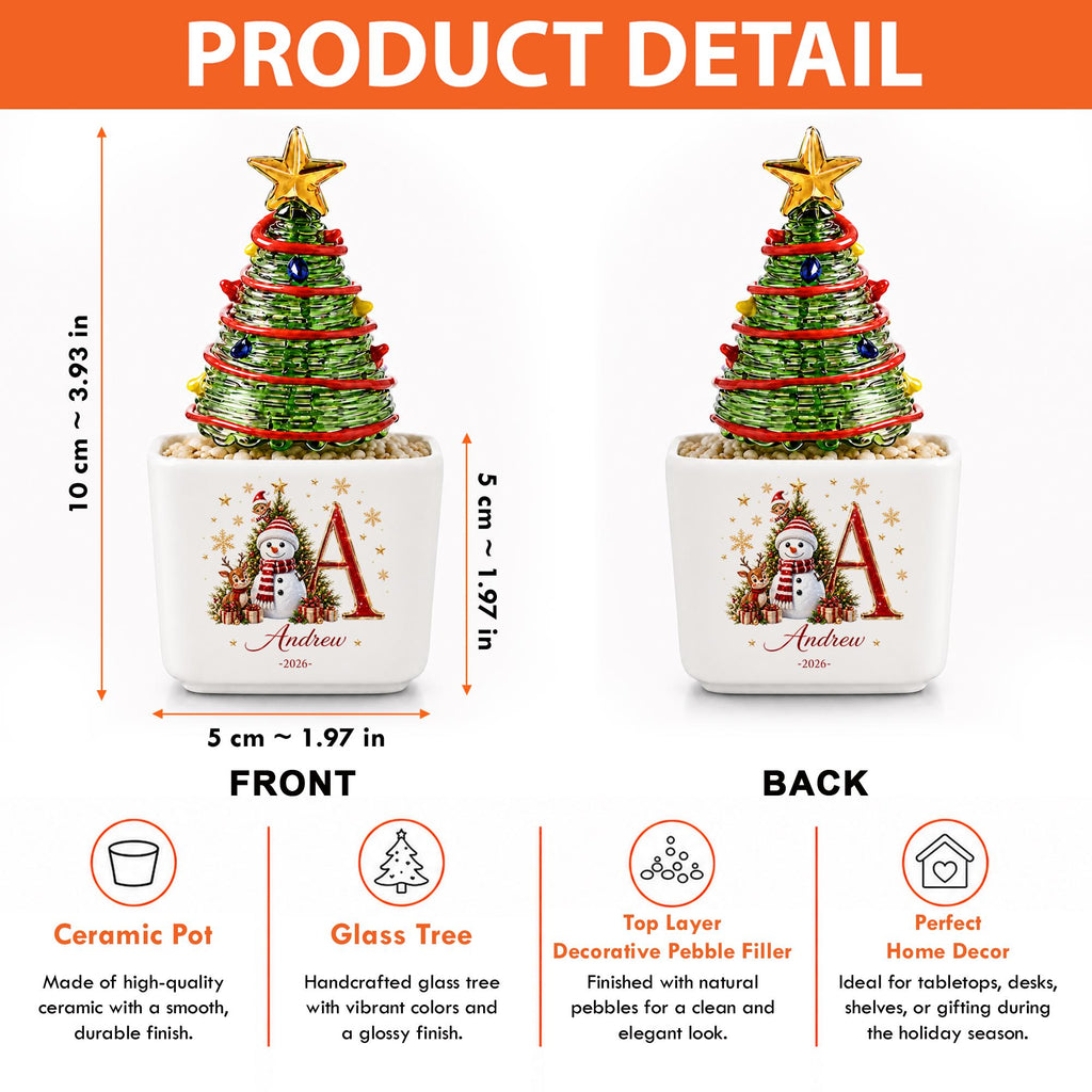 May include: A decorative glass Christmas tree with a gold star topper, red garland, and colorful ornaments, set in a white ceramic pot. The pot features a snowman graphic, the letter A, and the text Andrew 2026. Dimensions are 3.93 inches tall.