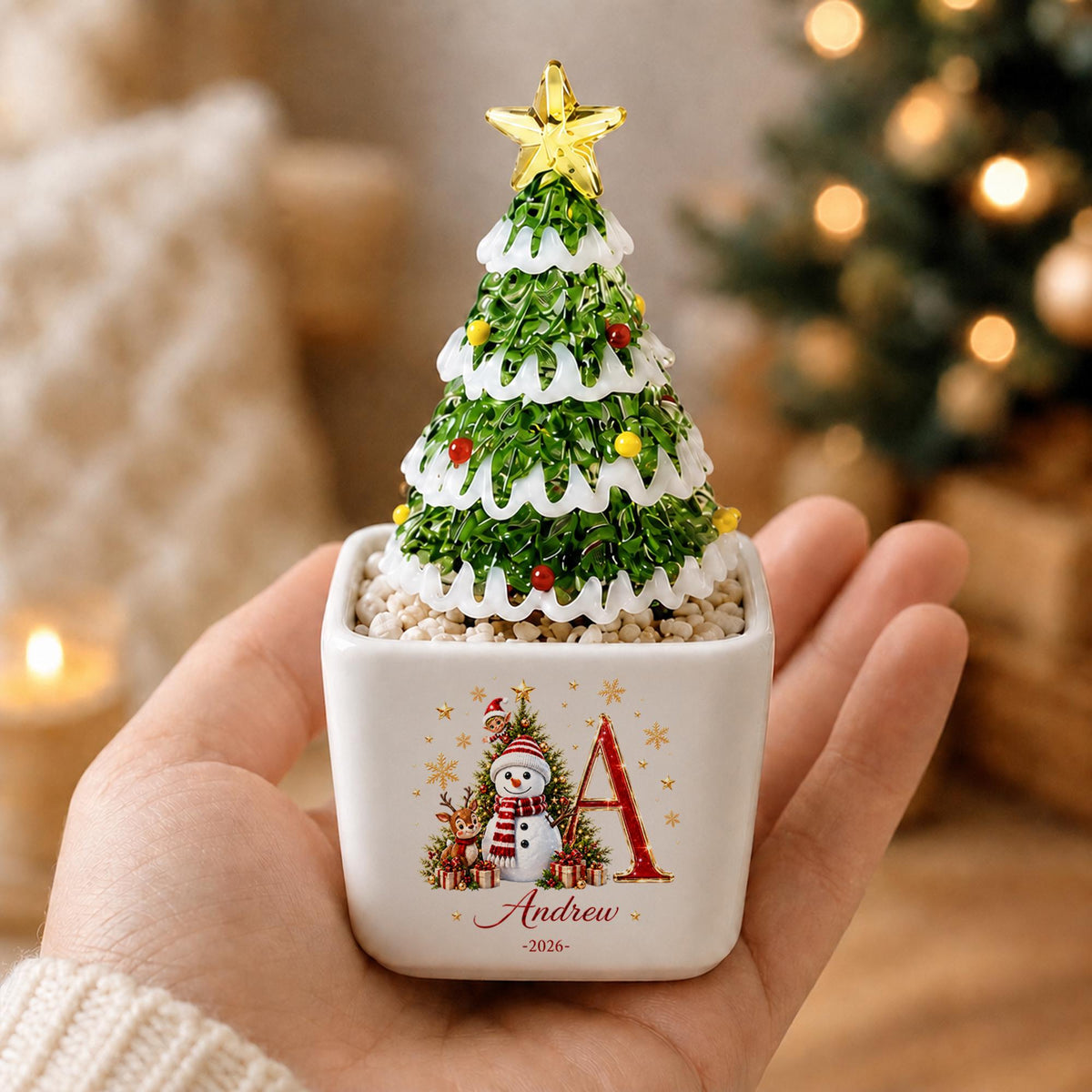 May include: A small decorative Christmas tree ornament in a white square ceramic pot. The tree is green with white snow-like trim, colorful baubles, and a yellow star topper. The pot features a graphic of a snowman, a reindeer, and the letter A.