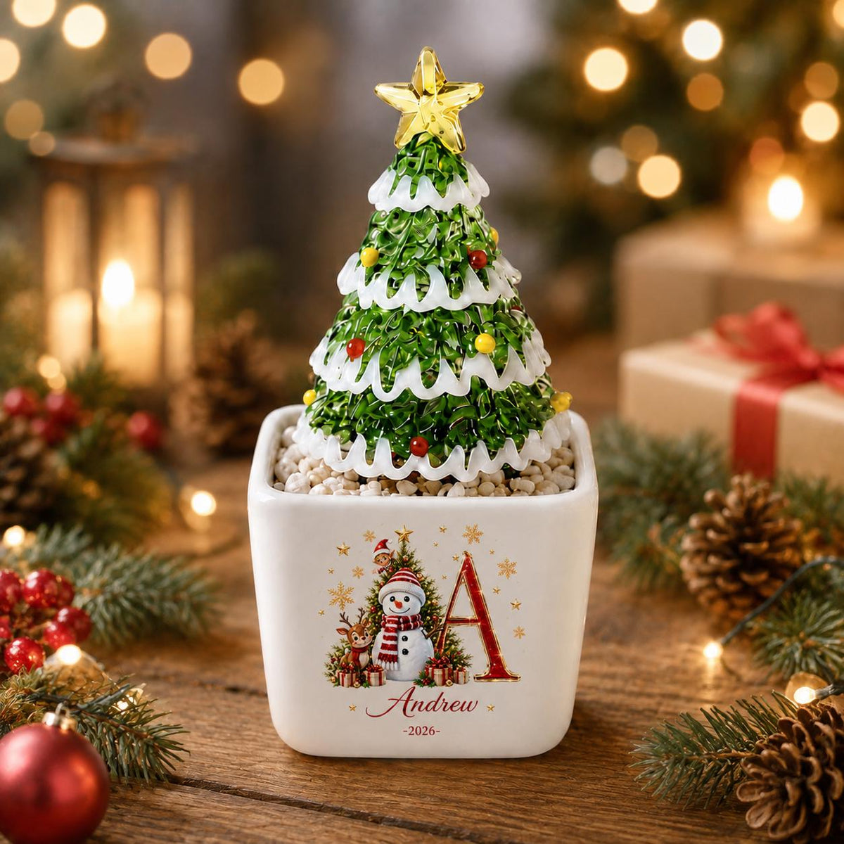 May include: A decorative tabletop Christmas tree with green foliage, white snow-like trim, and small colorful ornaments, topped with a yellow star. It sits in a white square ceramic planter featuring a snowman, a reindeer, and the name Andrew 2026.
