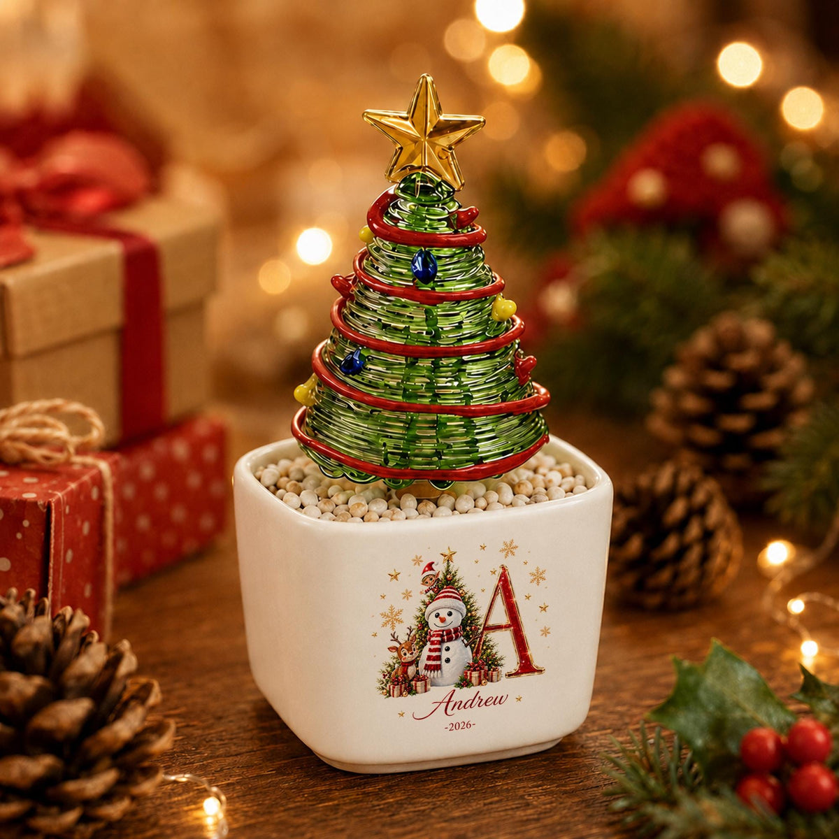 May include: A decorative tabletop Christmas tree made of green wire coils with a gold star topper, red garland, and small colored beads, set in a white square ceramic planter featuring a snowman graphic, the letter A, and the name Andrew 2026.