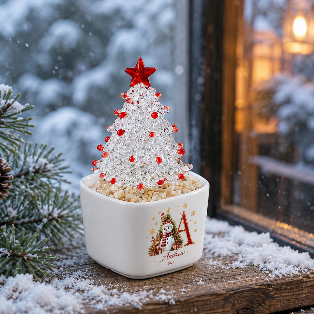 May include: A clear glass Christmas tree ornament with red glass beads and a red star topper, set in a white ceramic square planter filled with small stones. The planter features a graphic of a snowman, a tree, the letter A, and the name Andrew 2026.