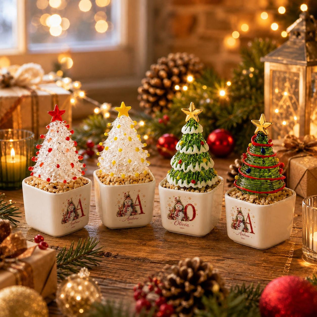 May include: Four miniature Christmas tree figurines in square white ceramic pots. Each tree features a unique design with a star topper and sits on a base of small pebbles. The pots are decorated with a snowman illustration and a large letter.