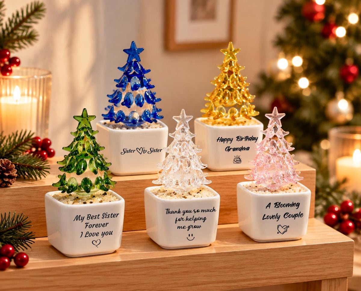 May include: Five miniature glass Christmas trees in white square pots. The trees are green, blue, clear, yellow, and pink. Each pot features black text: 'My Best Sister Forever I Love you', 'Sister ♡ Sister', 'Thank you so much for helping me grow', 'Happy Birthday Grandma', and 'A Blooming Lovely Couple'.
