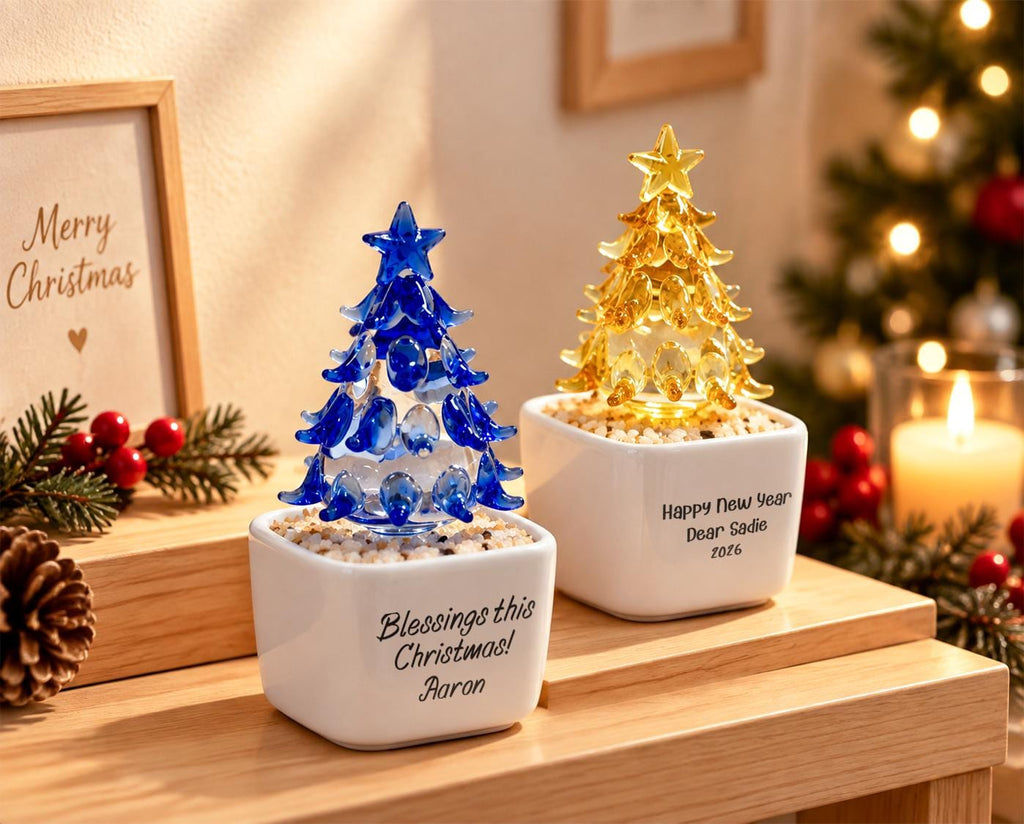 May include: Two glass Christmas tree ornaments in white square planters filled with small stones. One tree is blue, the other is yellow, both topped with stars. The blue planter reads Blessings this Christmas! Aaron, the yellow reads Happy New Year Dear Sadie 2026.