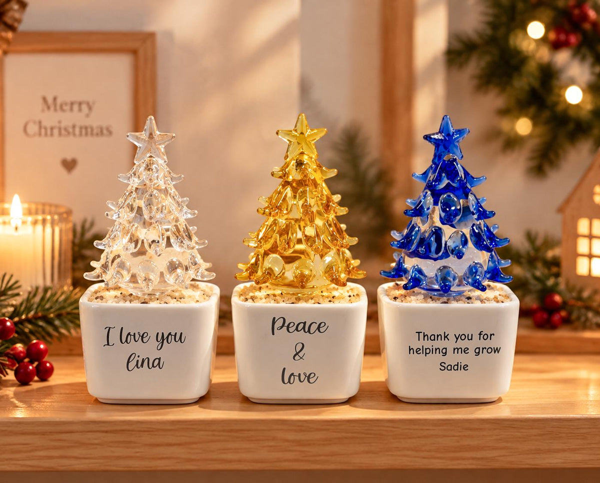 May include: Three glass Christmas tree ornaments in white square ceramic pots. The left pot says I love you Lina, the center says Peace & love, and the right says Thank you for helping me grow Sadie. The trees are clear, gold, and blue glass.
