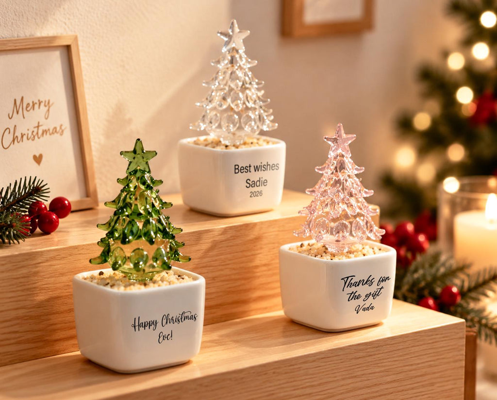 May include: Three glass Christmas tree ornaments in white square ceramic pots filled with small pebbles. One tree is green, one is clear, and one is pink. Each pot features black printed text: Happy Christmas Eve, Best wishes Sadie 2026, and Thanks for the gift Vada.