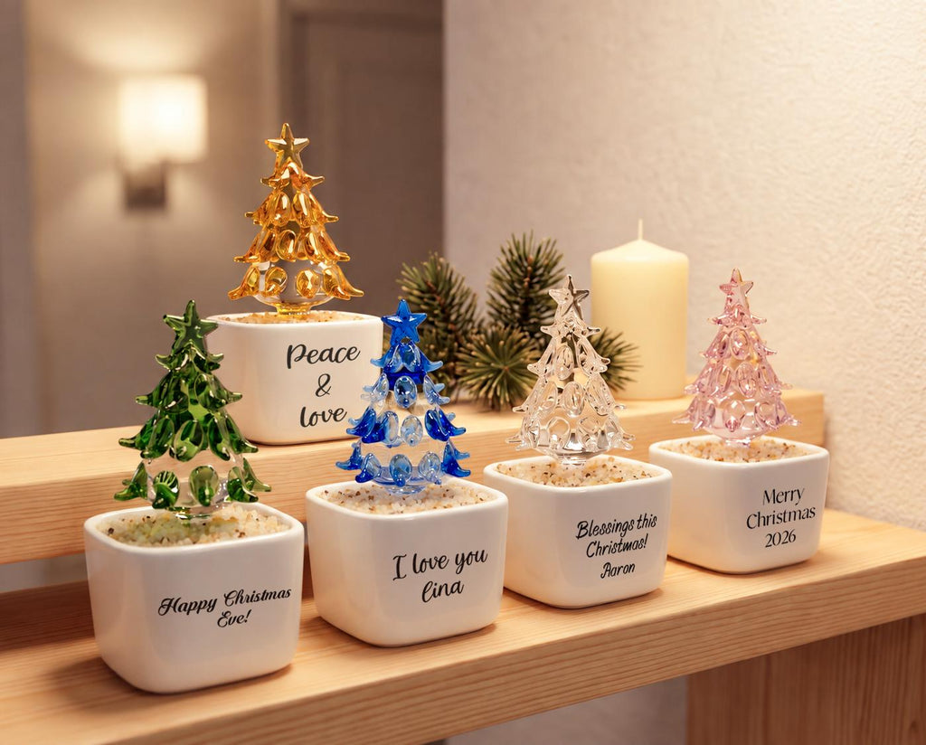 May include: Five glass Christmas tree ornaments in white square ceramic pots filled with small stones. The trees are gold, green, blue, clear, and pink. Each pot features black text: Peace & Love, Happy Christmas Eve!, I love you Lina, Blessings this Christmas! Aaron, and Merry Christmas 2026.
