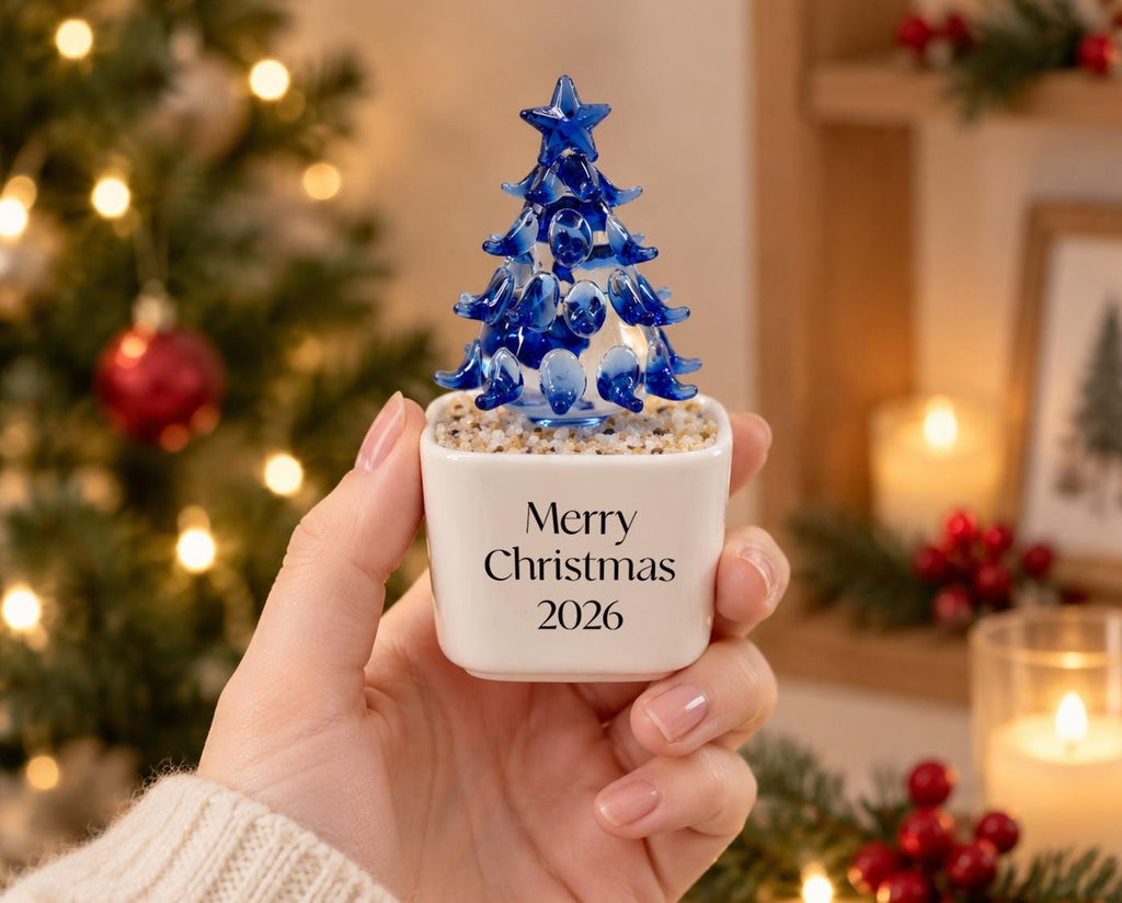May include: A small, white, square ceramic planter filled with light-colored pebbles and topped with a blue glass Christmas tree figurine. The front of the planter features the black printed text Merry Christmas 2026 in a simple serif font.