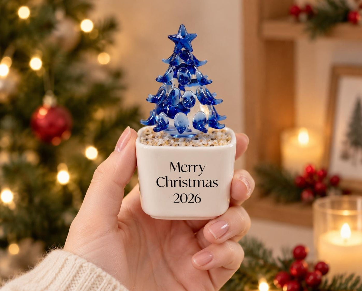 May include: A small, white, square ceramic planter filled with light-colored pebbles and topped with a blue glass Christmas tree figurine. The front of the planter features the black printed text Merry Christmas 2026 in a simple serif font.