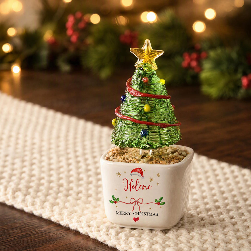 May include: A green wire Christmas tree ornament with a yellow star topper, colorful beads, and a red ribbon, set in a white square ceramic planter filled with small pebbles. The planter features the name Helene, a Santa hat, and text.