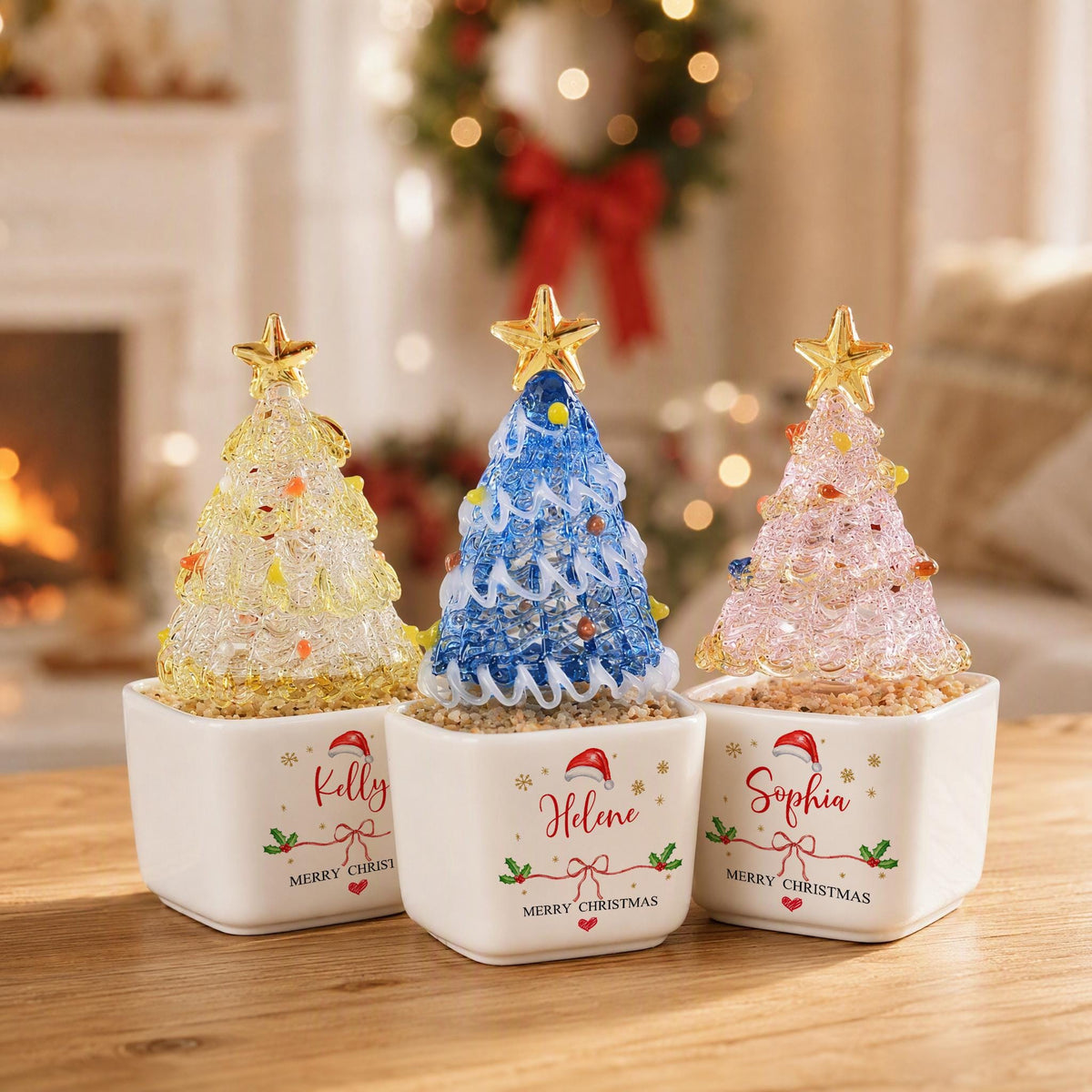 May include: Three decorative glass Christmas trees in white square ceramic pots. The trees are yellow, blue, and pink with gold stars on top. Each pot features a name, Kelly, Helene, or Sophia, with a Santa hat, holly, and Merry Christmas text.