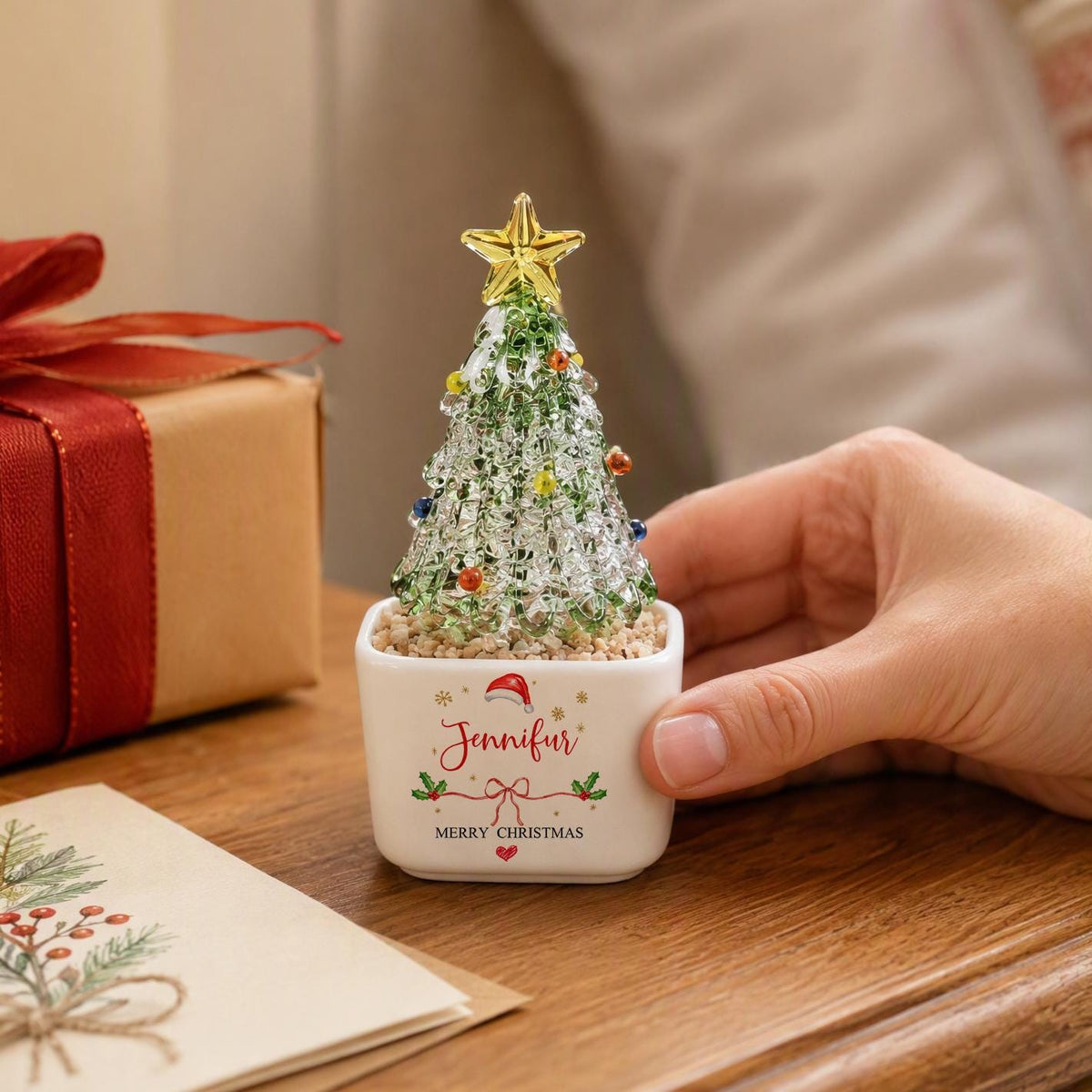 May include: A miniature glass Christmas tree with green and clear textured branches, topped with a yellow star and small colorful ornaments. It sits in a white square ceramic planter featuring the name Jennifer, a Santa hat, and text.