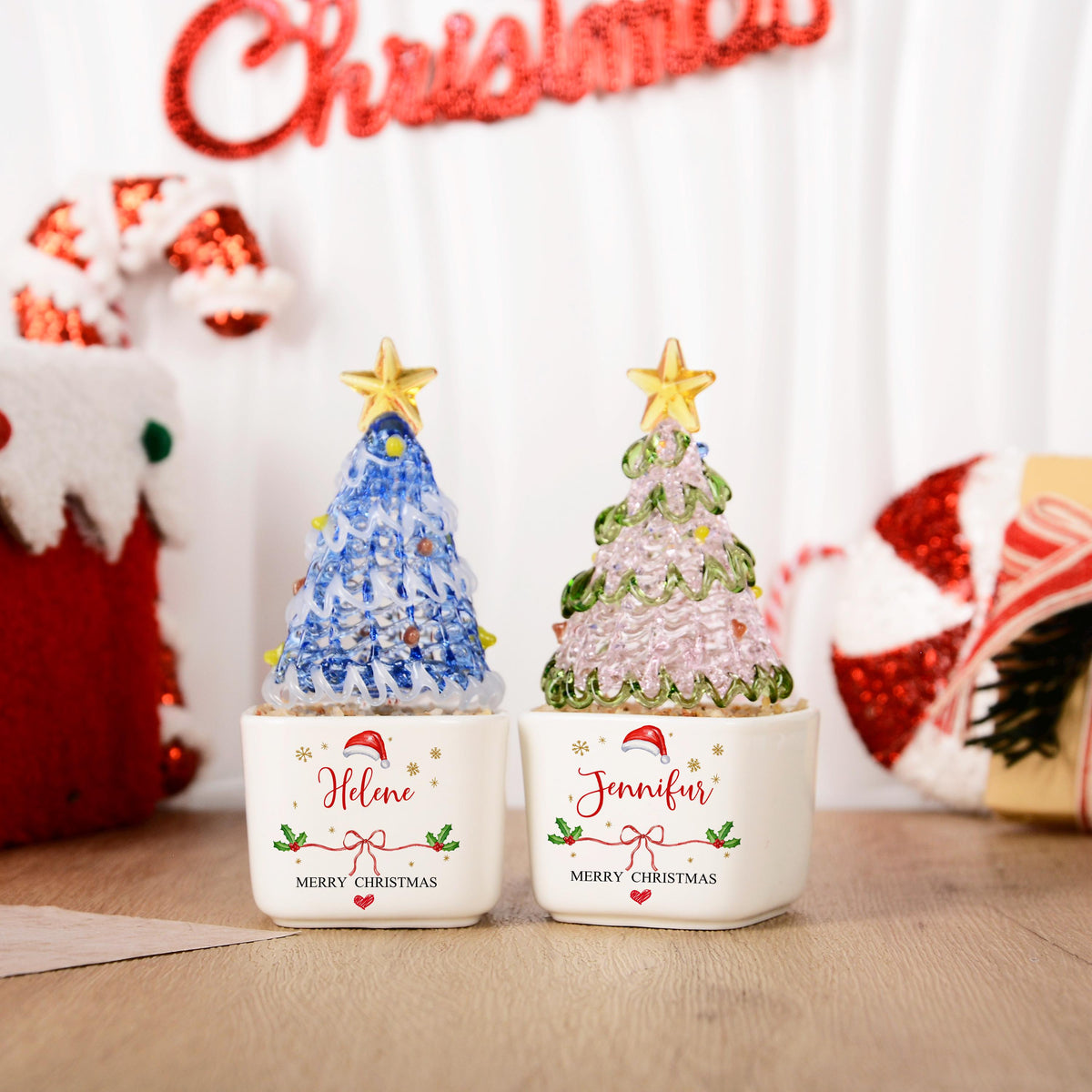 May include: Two glass Christmas tree figurines in white square ceramic pots. The left tree is blue with a yellow star, and the right tree is green with a yellow star. The pots feature the names Helene and Jennifur with Merry Christmas text.