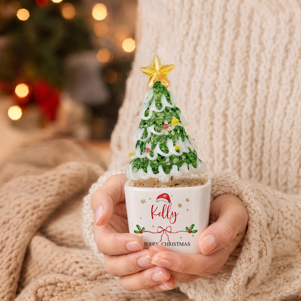 May include: A glass Christmas tree figurine with green branches, white snow-like accents, and a yellow star topper. The tree sits in a white square ceramic pot featuring the name Kelly, a red Santa hat, and the text Merry Christmas.