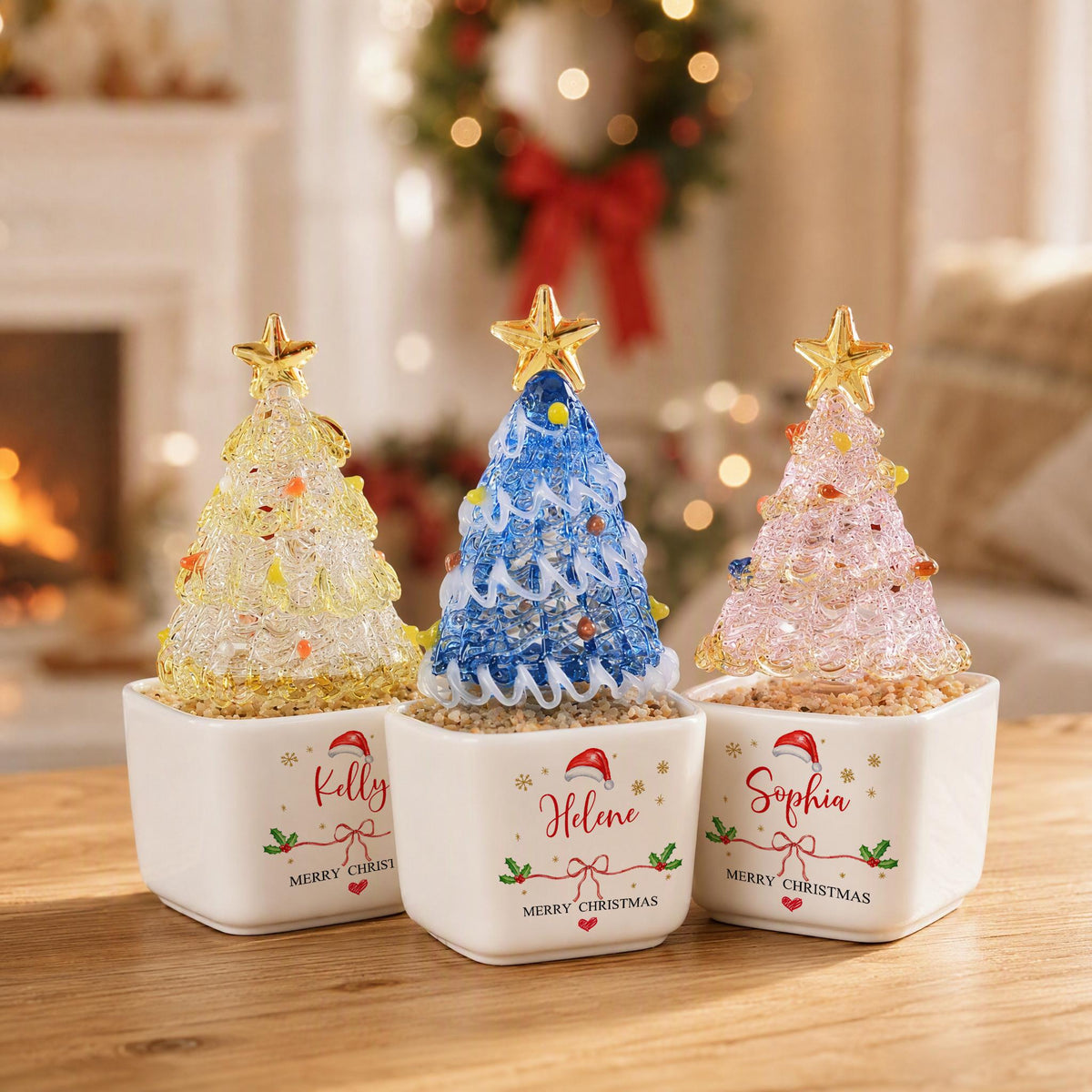 May include: Three decorative glass Christmas trees in white square ceramic pots. The trees are yellow, blue, and pink with gold stars on top. Each pot features a name, Kelly, Helene, or Sophia, with a Santa hat, holly, and Merry Christmas text.