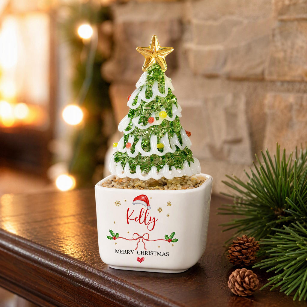 May include: A glass Christmas tree figurine with green textured foliage, white swirled accents, and a gold star topper. It sits in a white square ceramic planter featuring a red Santa hat, the name Kelly, and the text Merry Christmas.