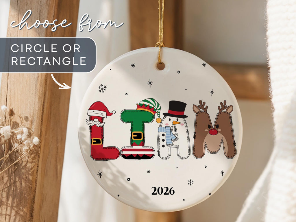 May include: A round white ceramic Christmas ornament featuring the name LIAM spelled out in festive character-themed letters: a Santa L, an elf I, a snowman A, and a reindeer M. The ornament includes the year 2026 and small black snowflake accents.