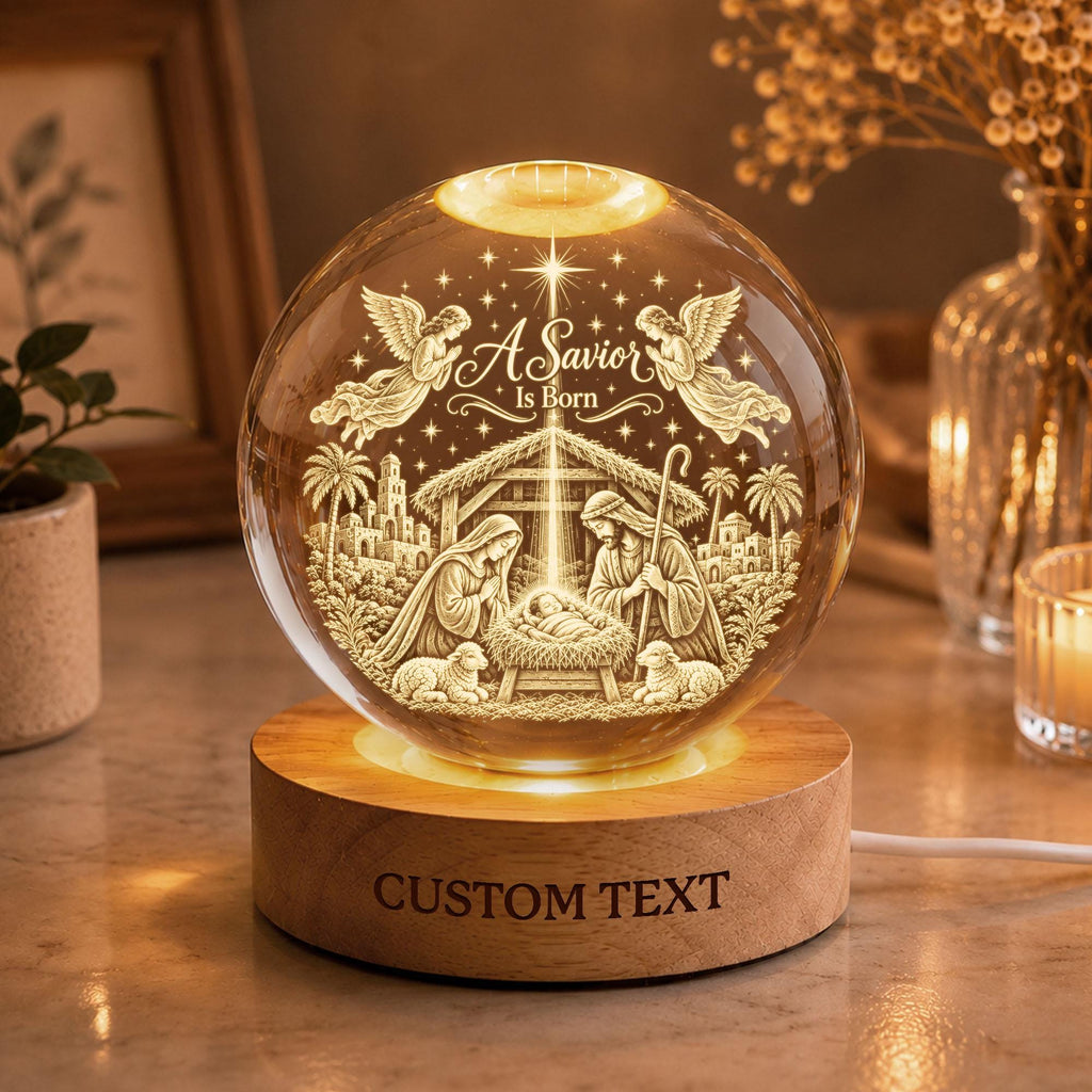 May include: A clear glass sphere lamp featuring an internal 3D laser-engraved nativity scene with angels, palm trees, and the text A Savior Is Born. The sphere rests on a round wooden base with the engraved text CUSTOM TEXT on the front side.