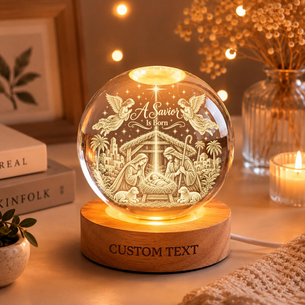 May include: A clear glass sphere night light featuring an etched nativity scene with the text A Savior Is Born. The sphere rests on a round wooden base with the words CUSTOM TEXT engraved on the front. The scene includes angels, palm trees, and figures.