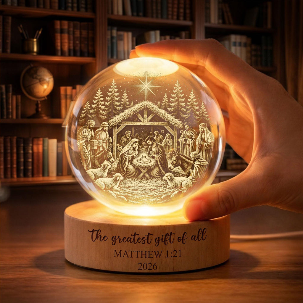May include: A clear glass sphere featuring an internal laser-etched nativity scene with a star, figures, and animals. The sphere rests on a cylindrical wooden base engraved with the text 'The greatest gift of all', 'MATTHEW 1:21', and '2026'.