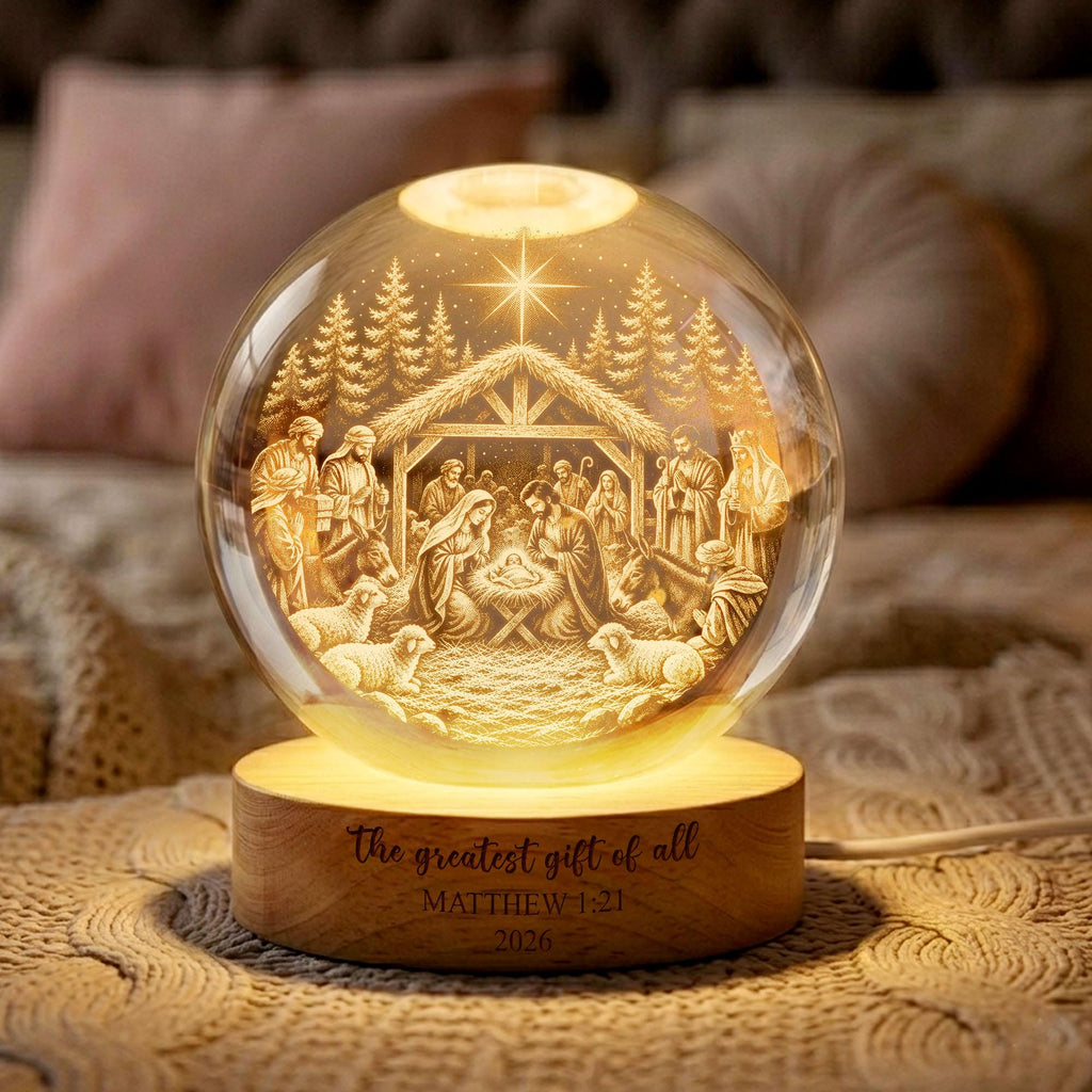 May include: A spherical glass night light featuring an etched nativity scene with a star, stable, and figures. The light sits on a round wooden base engraved with the text: The greatest gift of all, MATTHEW 1:21, 2026. It emits a warm, golden glow.