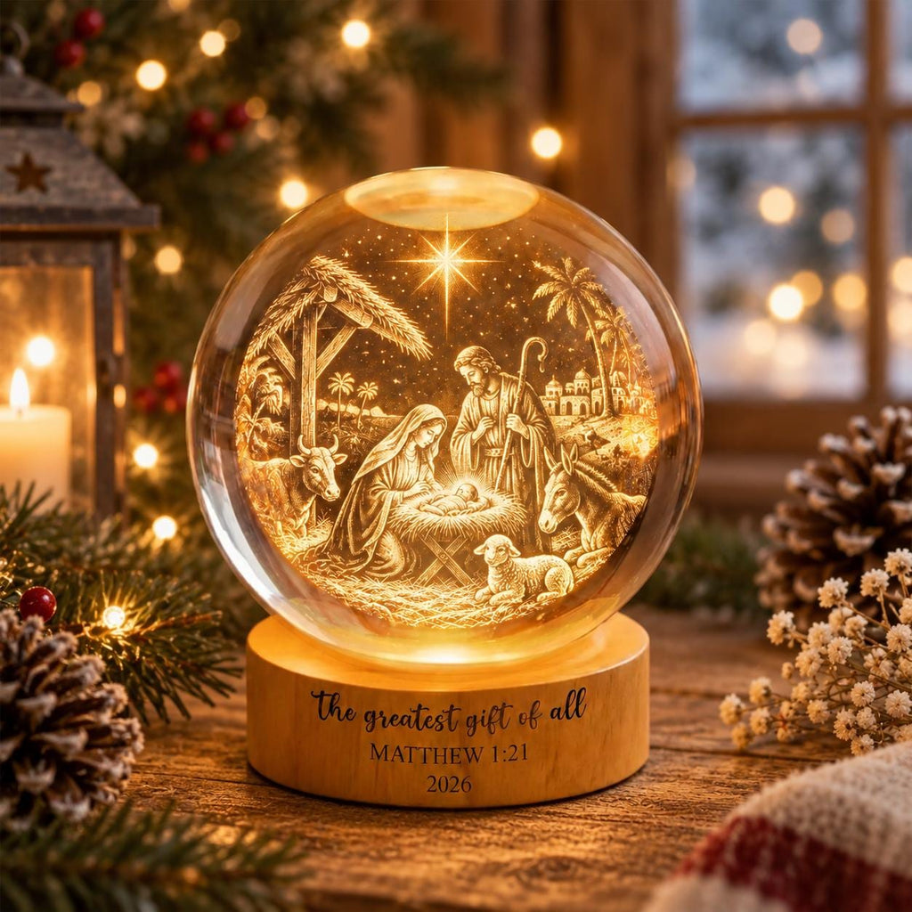 May include: A spherical glass ornament featuring an illuminated nativity scene with figures of Mary, Joseph, and the infant Jesus, along with a cow, donkey, and lamb. The wooden base is engraved with the text The greatest gift of all MATTHEW 1:21 2026.