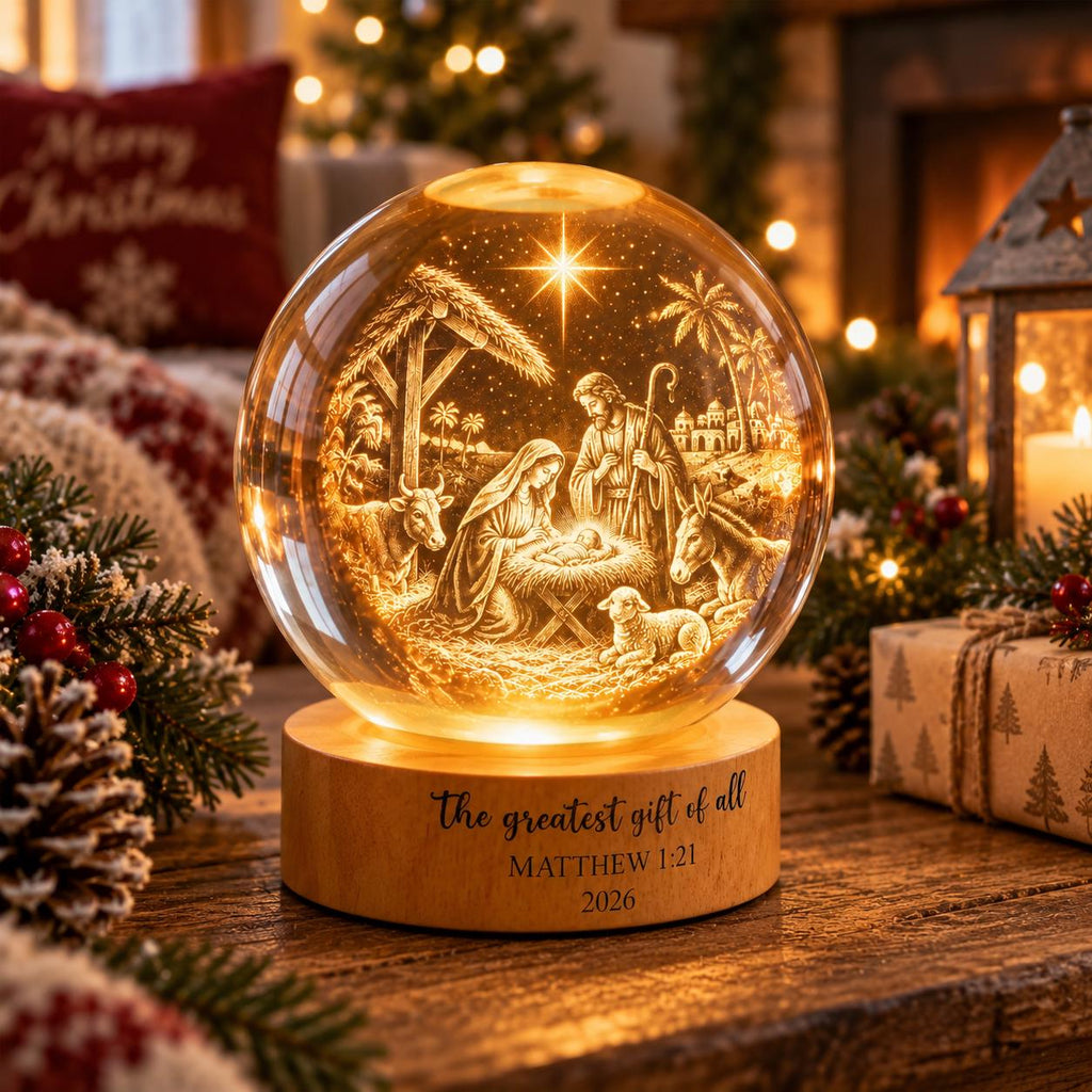 May include: A spherical glass ornament featuring an illuminated nativity scene with figures of Mary, Joseph, and the infant Jesus. The wooden base is engraved with the text: The greatest gift of all, MATTHEW 1:21, 2026. The scene includes a star.