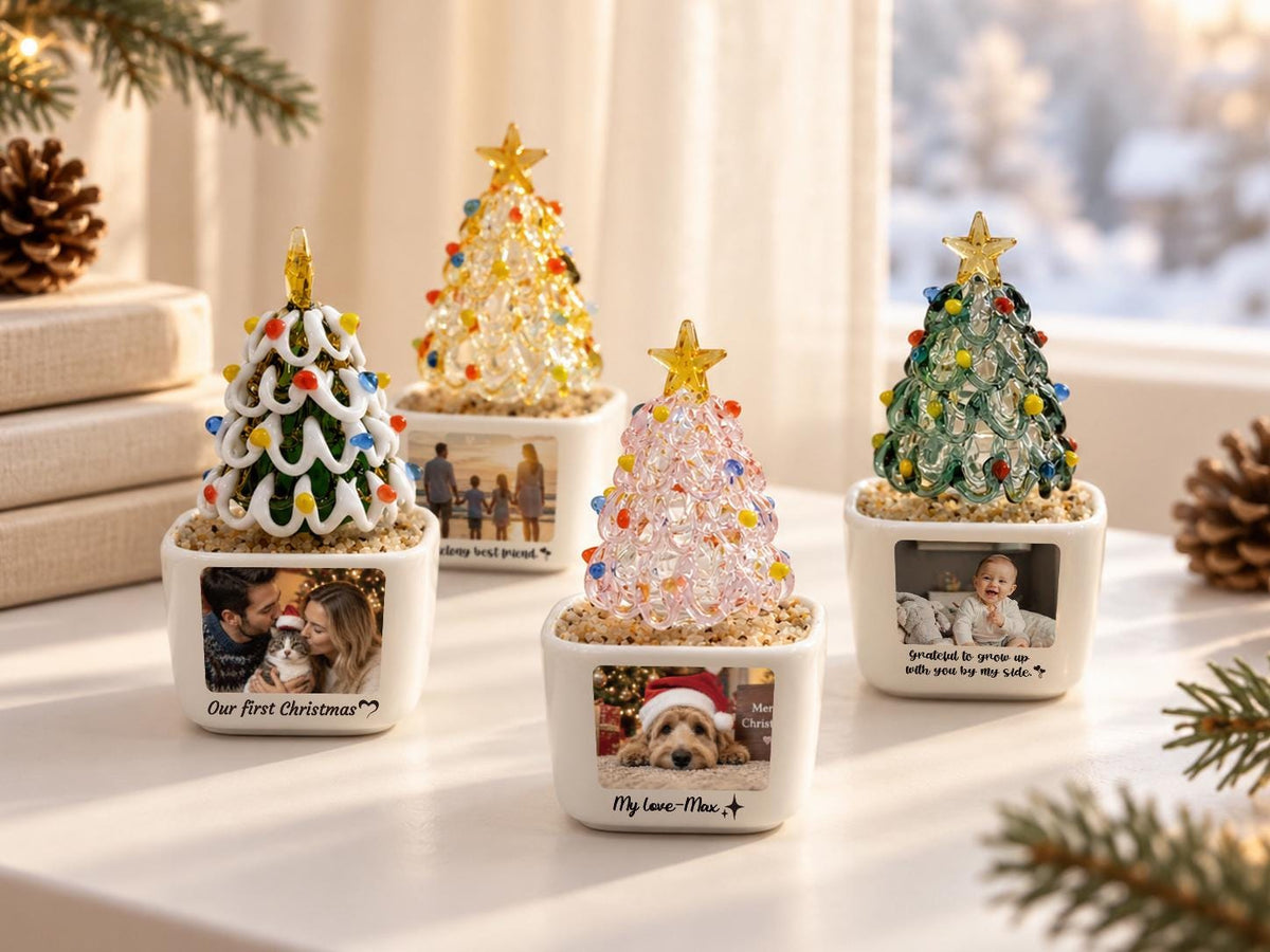 May include: Four decorative tabletop Christmas trees in white square ceramic planters. Each tree features a glass structure with colorful lights and a gold star topper. The planters display personalized photographs and text including Our first Christmas.