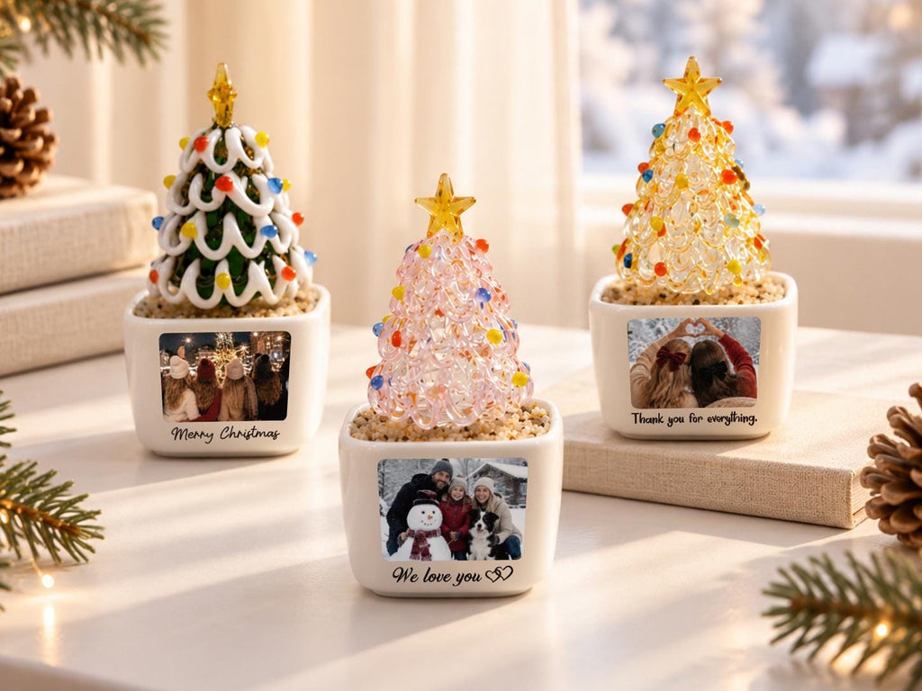 May include: Three miniature glass Christmas trees in white square ceramic pots. The left tree is green with white swirls and colorful beads. The center tree is pink with beads. The right tree is clear with beads. Each pot features a photo and text.