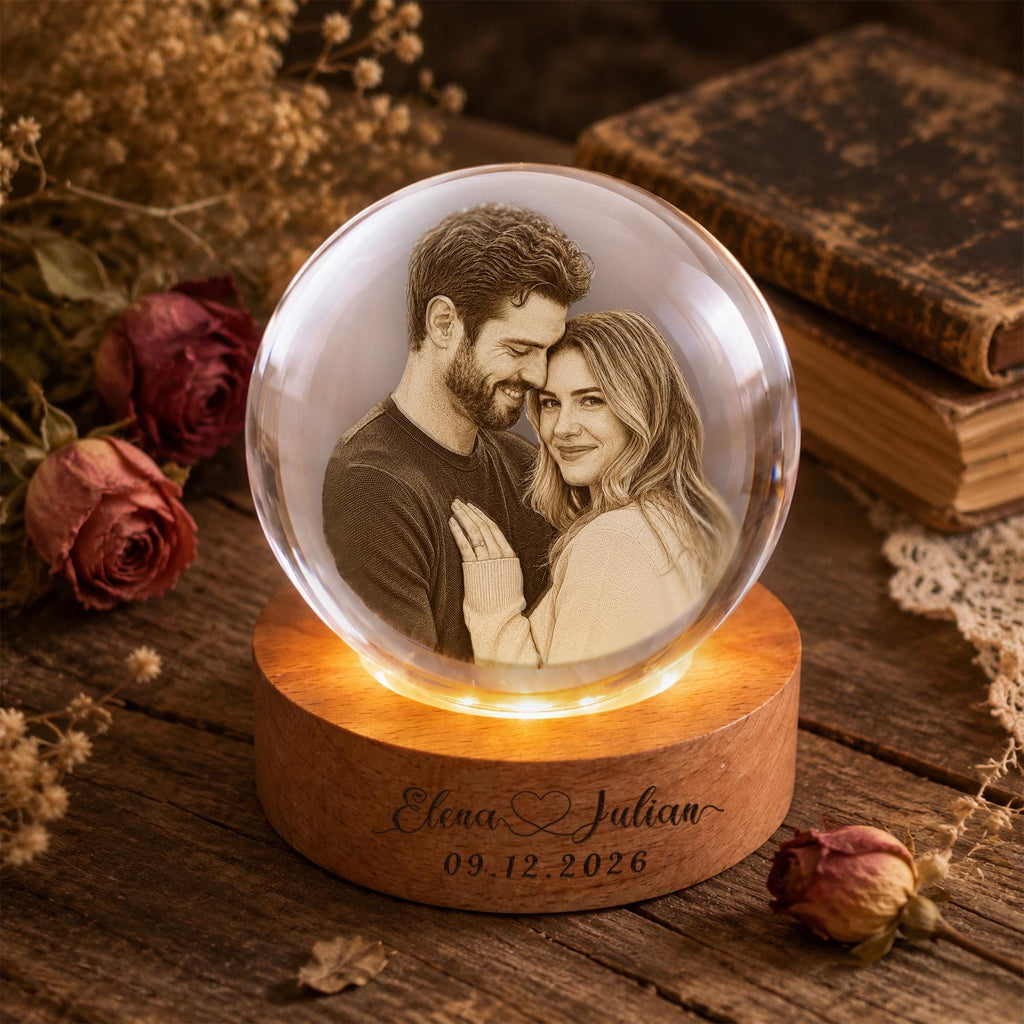May include: A clear glass sphere containing a laser-etched portrait of a couple, mounted on a circular wooden base. The base features the engraved text Elena & Julian and the date 09.12.2026, with internal lighting illuminating the glass sphere.