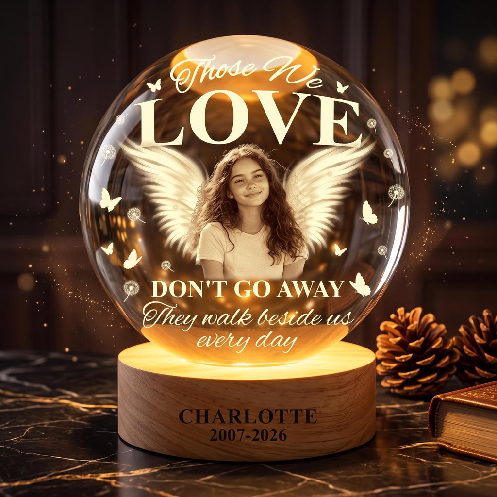 May include: A spherical glass memorial lamp on a cylindrical wooden base. The glass features a portrait of a young person with angel wings, butterflies, and dandelions. Text reads: Those We LOVE DON'T GO AWAY They walk beside us every day. CHARLOTTE 2007-2026.