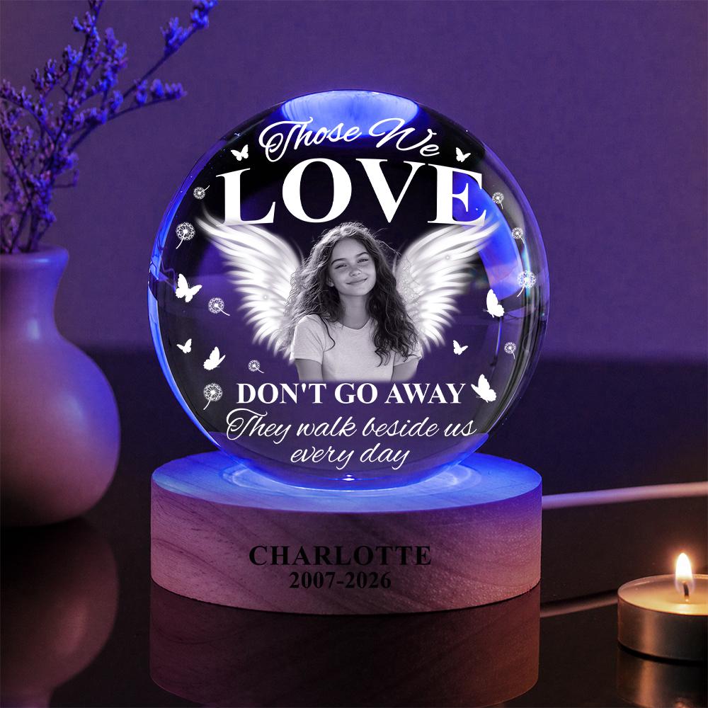 May include: A spherical glass memorial ornament featuring a black and white portrait of a person with angel wings. The text reads: Those We LOVE DON'T GO AWAY They walk beside us every day. The wooden base is engraved with CHARLOTTE 2007-2026.