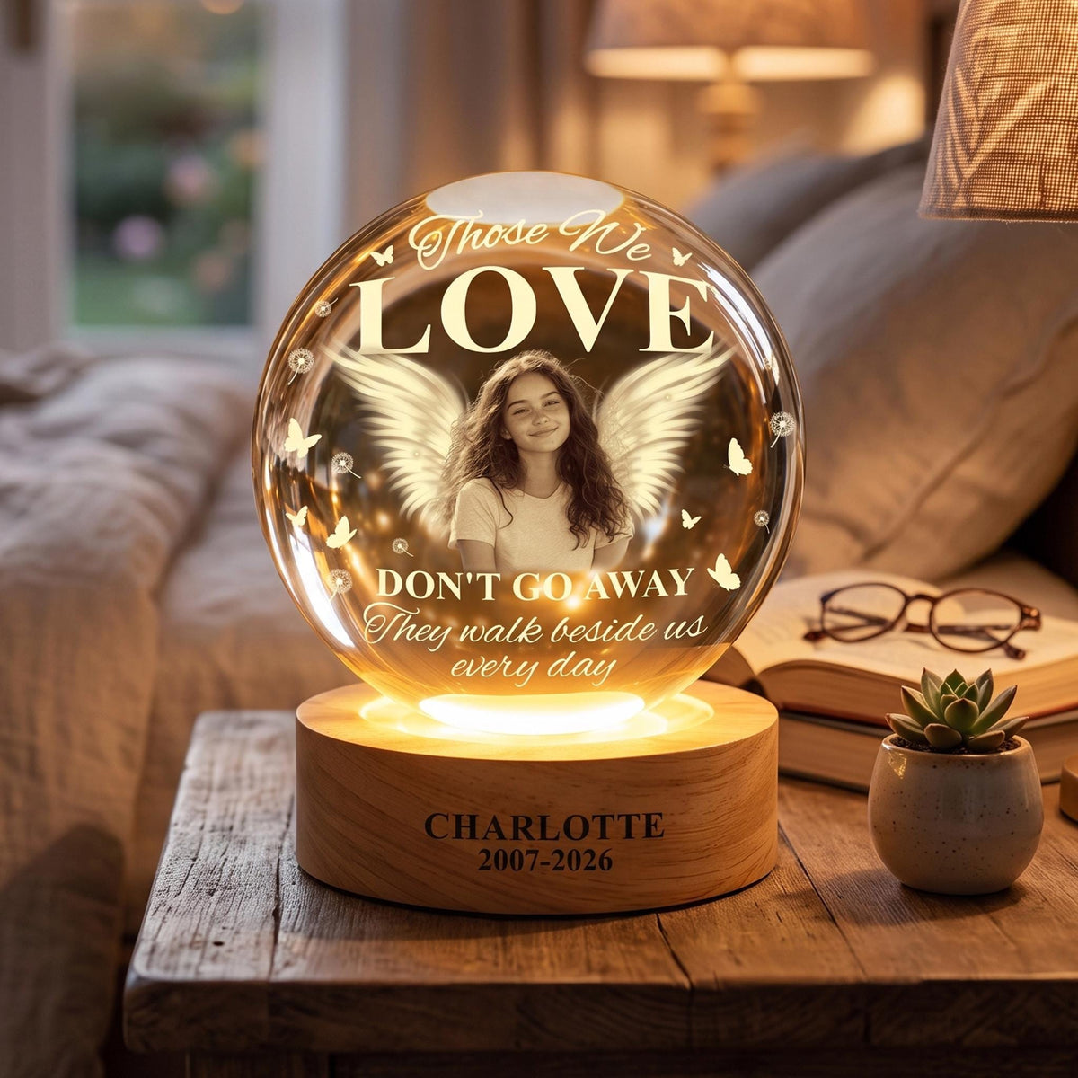 May include: A spherical glass memorial lamp featuring a photo of a person with angel wings, butterflies, and the text "Those We LOVE DON'T GO AWAY They walk beside us every day". The wooden base is engraved with "CHARLOTTE 2007-2026".