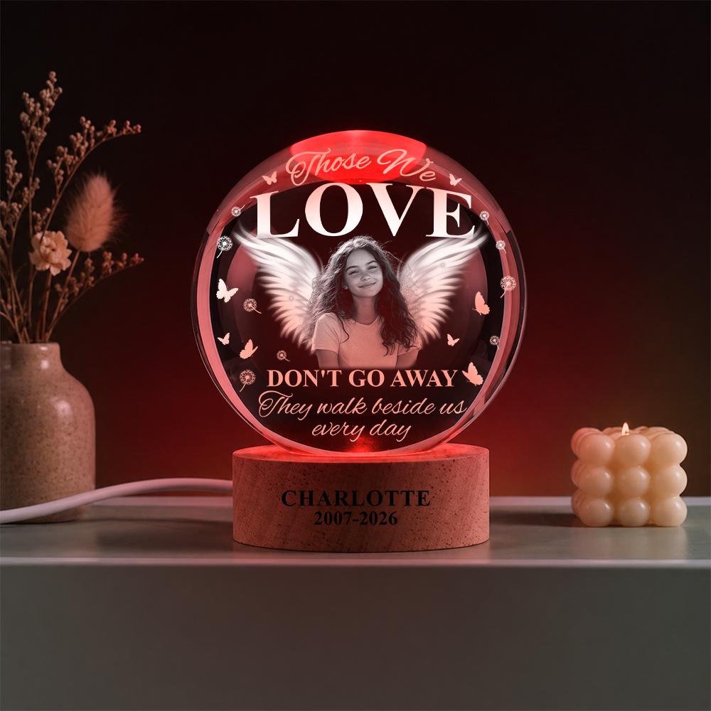 May include: A circular glass memorial lamp with a red glow, featuring a black and white portrait of a person with angel wings. The text reads: Those We LOVE DON'T GO AWAY They walk beside us every day. The wooden base is engraved: CHARLOTTE 2007-2026.