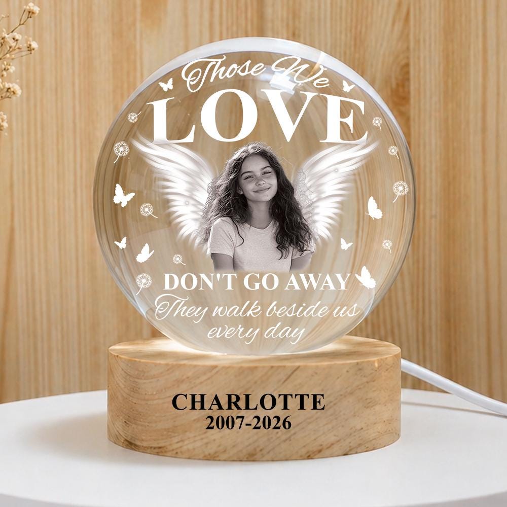 May include: A circular glass plaque on a light wood base featuring a black and white portrait of a person with angel wings. The text reads: Those We LOVE DON'T GO AWAY They walk beside us every day. The base is engraved with CHARLOTTE 2007-2026.