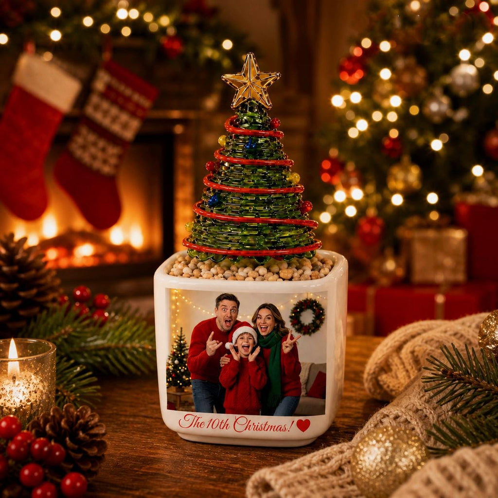May include: A square white ceramic planter featuring a printed photograph of a family of three wearing red sweaters, with the text "The 10th Christmas!" and a red heart. A green wire Christmas tree with a gold star topper sits in the planter.