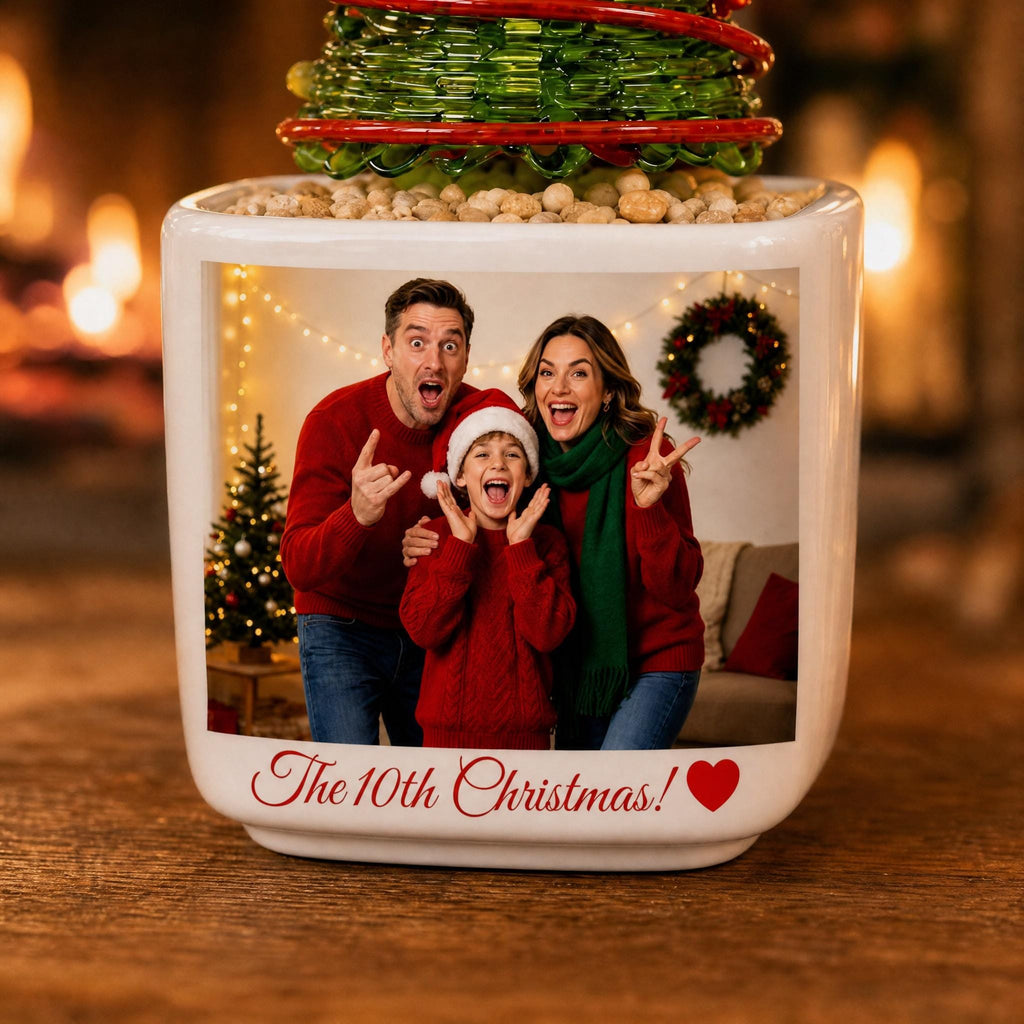 May include: A white ceramic planter featuring a printed photograph of a family of three wearing red sweaters and a green scarf, with the text "The 10th Christmas!" and a red heart icon. The planter is topped with a green glass decorative tree.